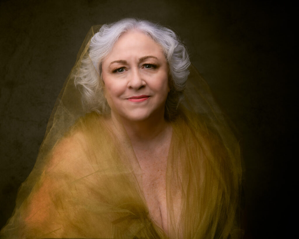 A beautiful woman with white hair, staring into the camera, with a wreath of gold tulle over her head.