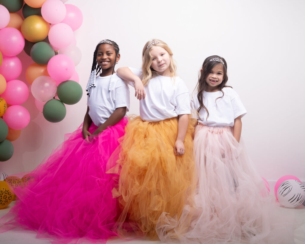 Three young girls in poufy tulle skirts and tiaras standing in front of balloons on a white wall.